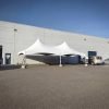 White high peak marquee tent with concrete ballasts set up in a Coquitlam warehouse parking lot beside loading bay doors and parked vehicles